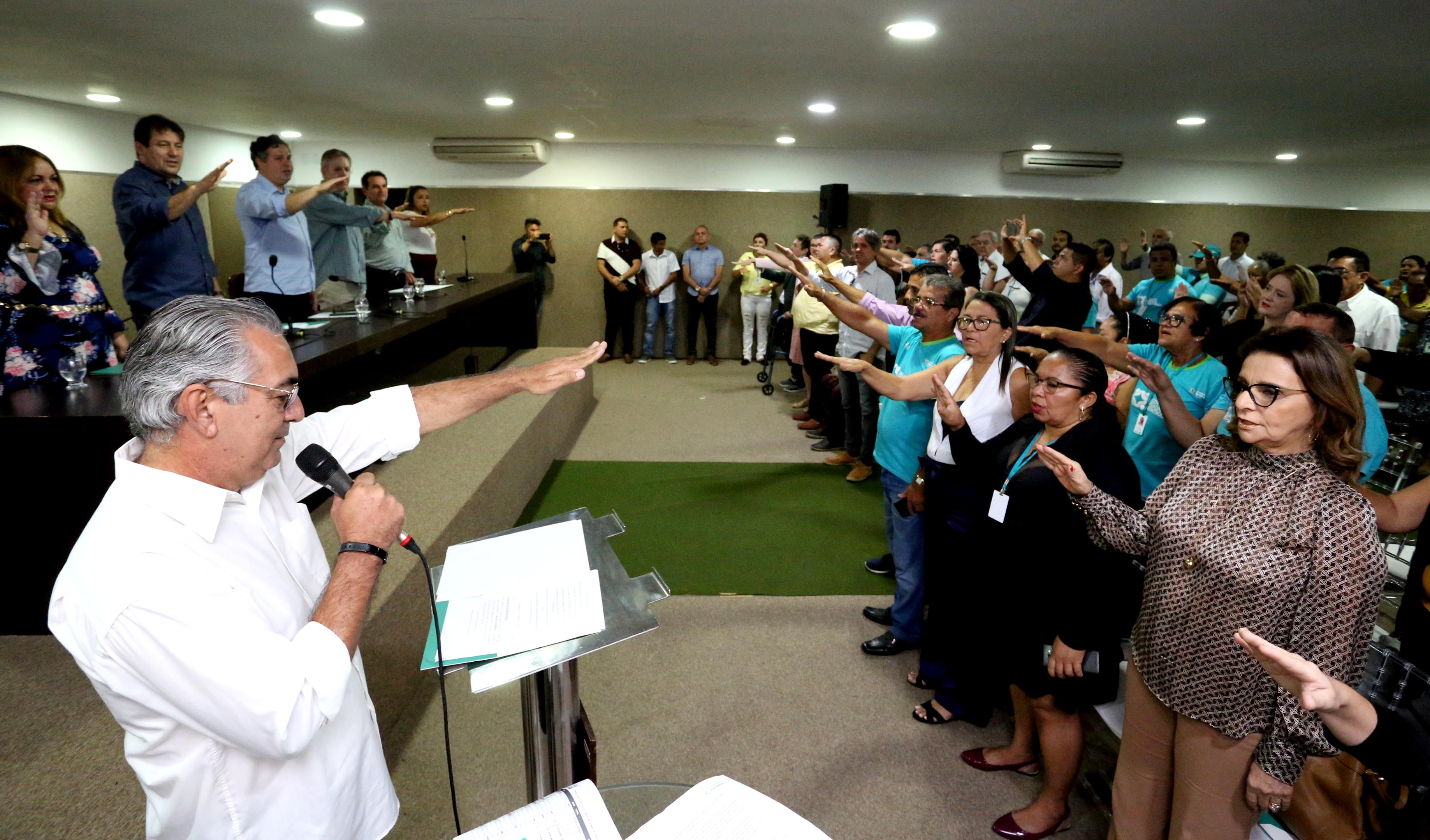 pessoas em auditório em pé em posição de juramento com braço extendido na frente corpo e homem falando em microfone